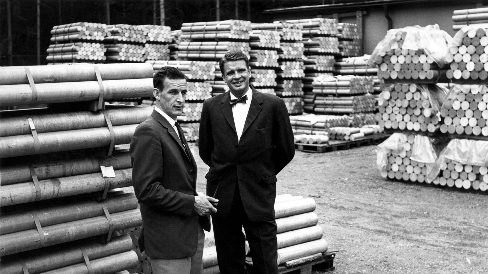 a couple of men standing in front of stacks of wood